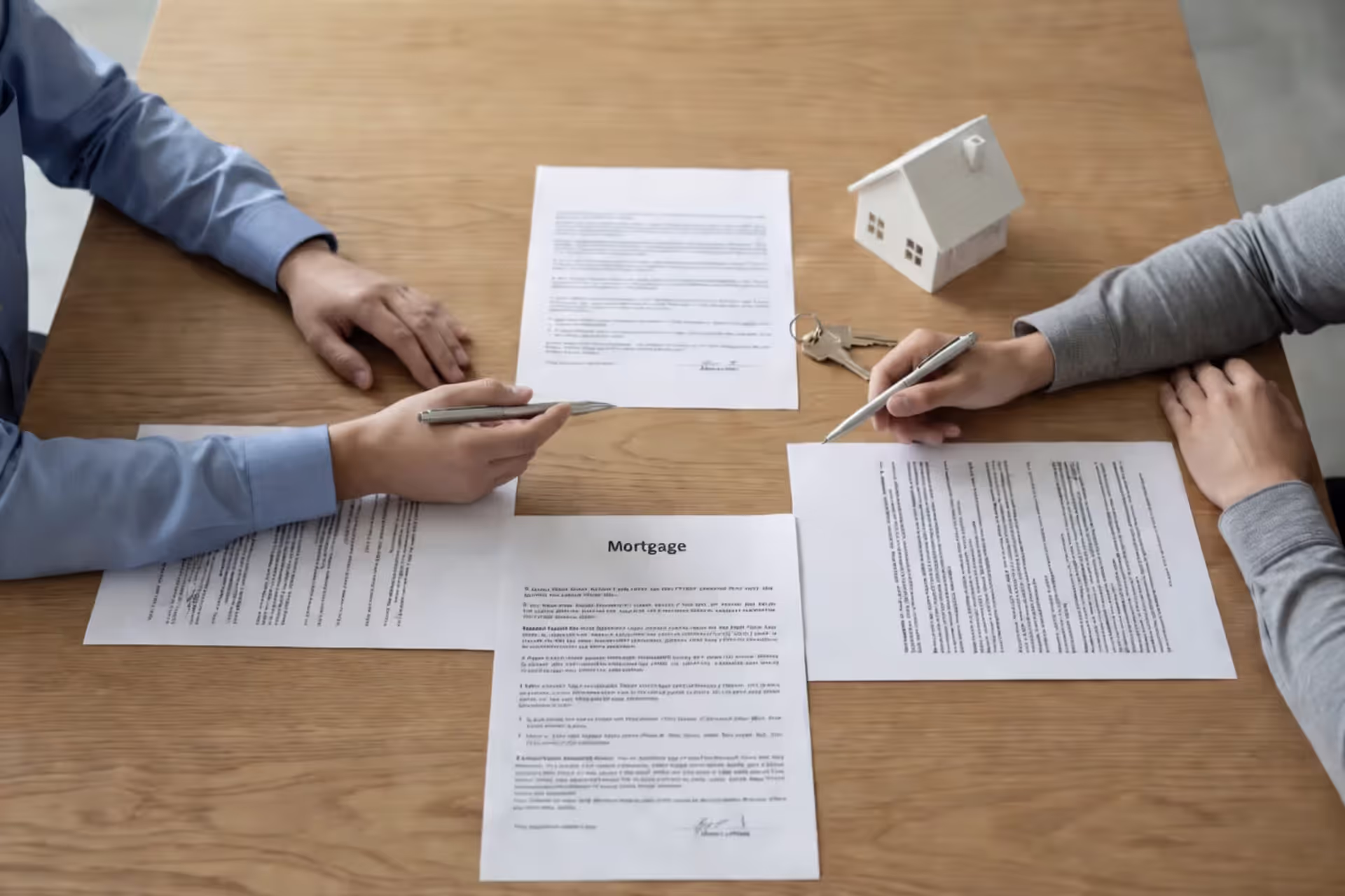 Top view of a closing table with mortgage documents, house keys, and a small house model, with two pairs of hands during a home purchase signing